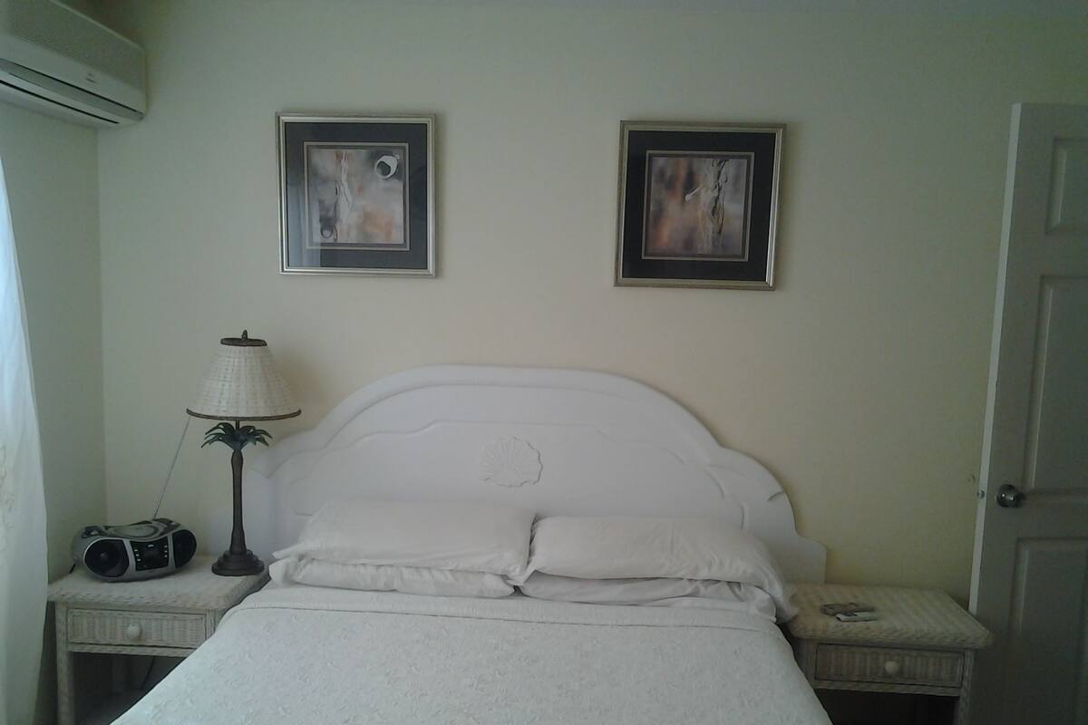 A neatly made bed with pillows, a lamp, and a radio on a bedside table. There are two framed pictures on the wall above the bed.