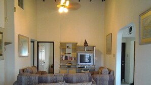 A living room with a large sectional sofa, a television, and a bookshelf. - Sungold House Barbados (Speightstown)