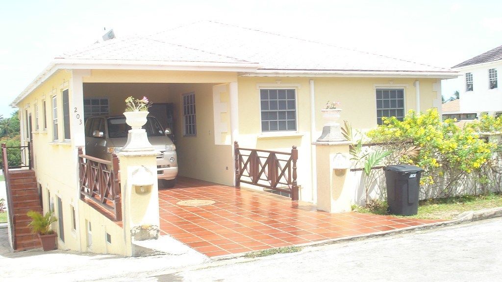 A two-story house with a tiled porch, a car parked in the driveway, and a trash bin to the side.