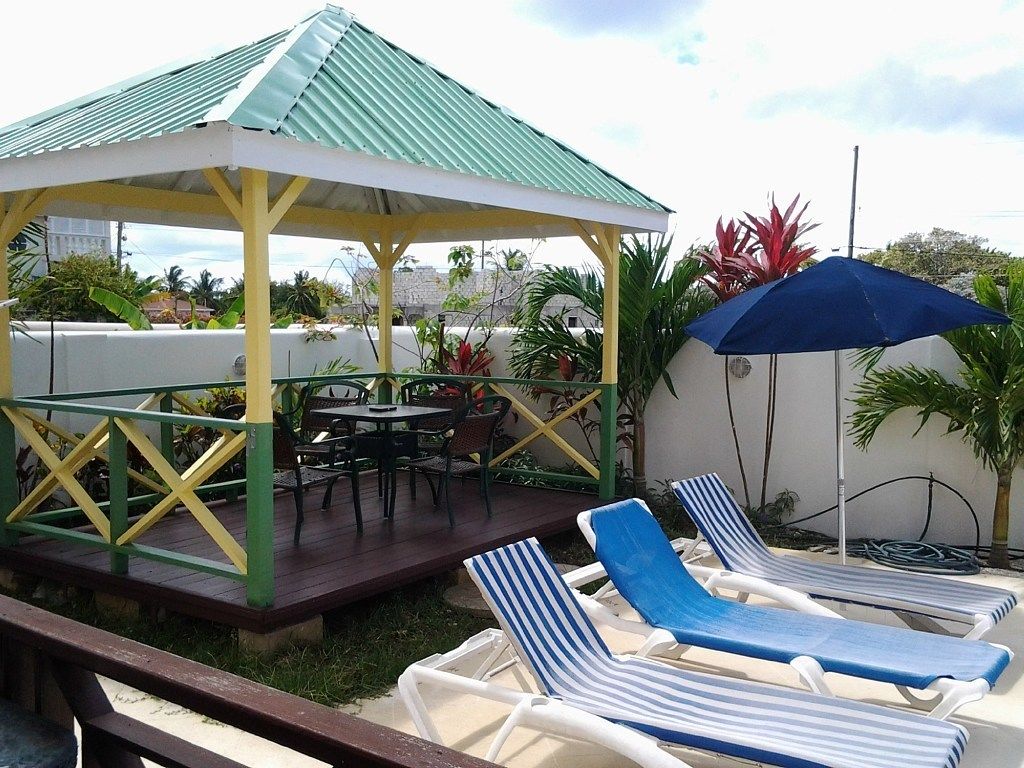 Un espacio al aire libre cubierto con sillones de descanso y una mesa, rodeado de plantas y un pabellón.