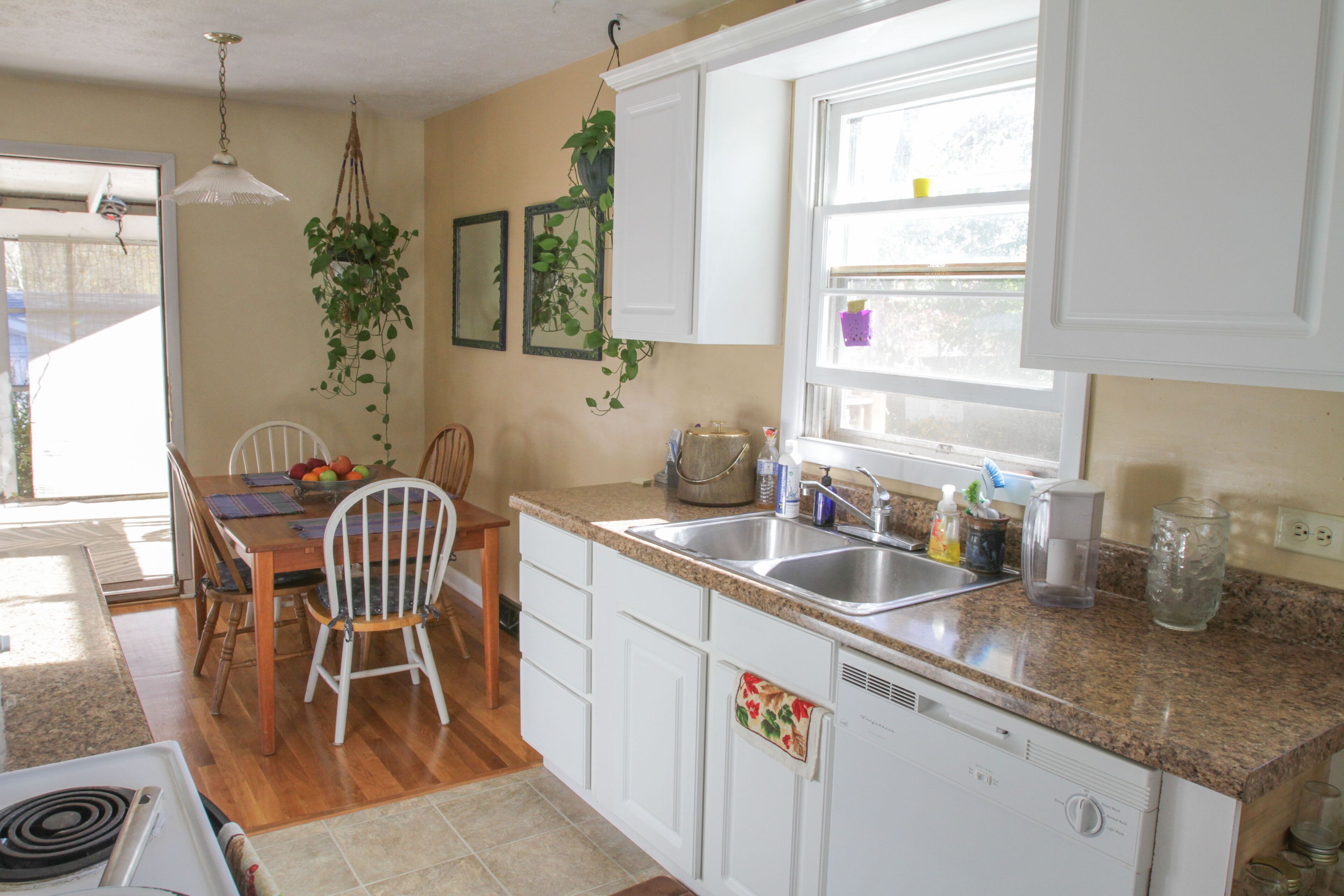 A kitchen with white cabinets, a granite countertop, a dining table with chairs, a hanging plant, and a window with a view.