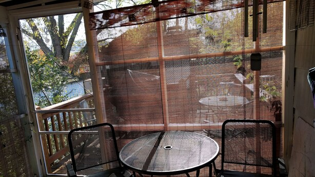 En overdækket terrasse med to metalstole og et rundt bord, med udsigt til udendørsområdet gennem et netvindue.