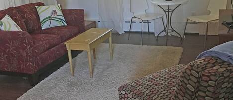 A living room with a red sofa, a wooden coffee table, a patterned armchair, a small white table with a flower vase, and a chair.