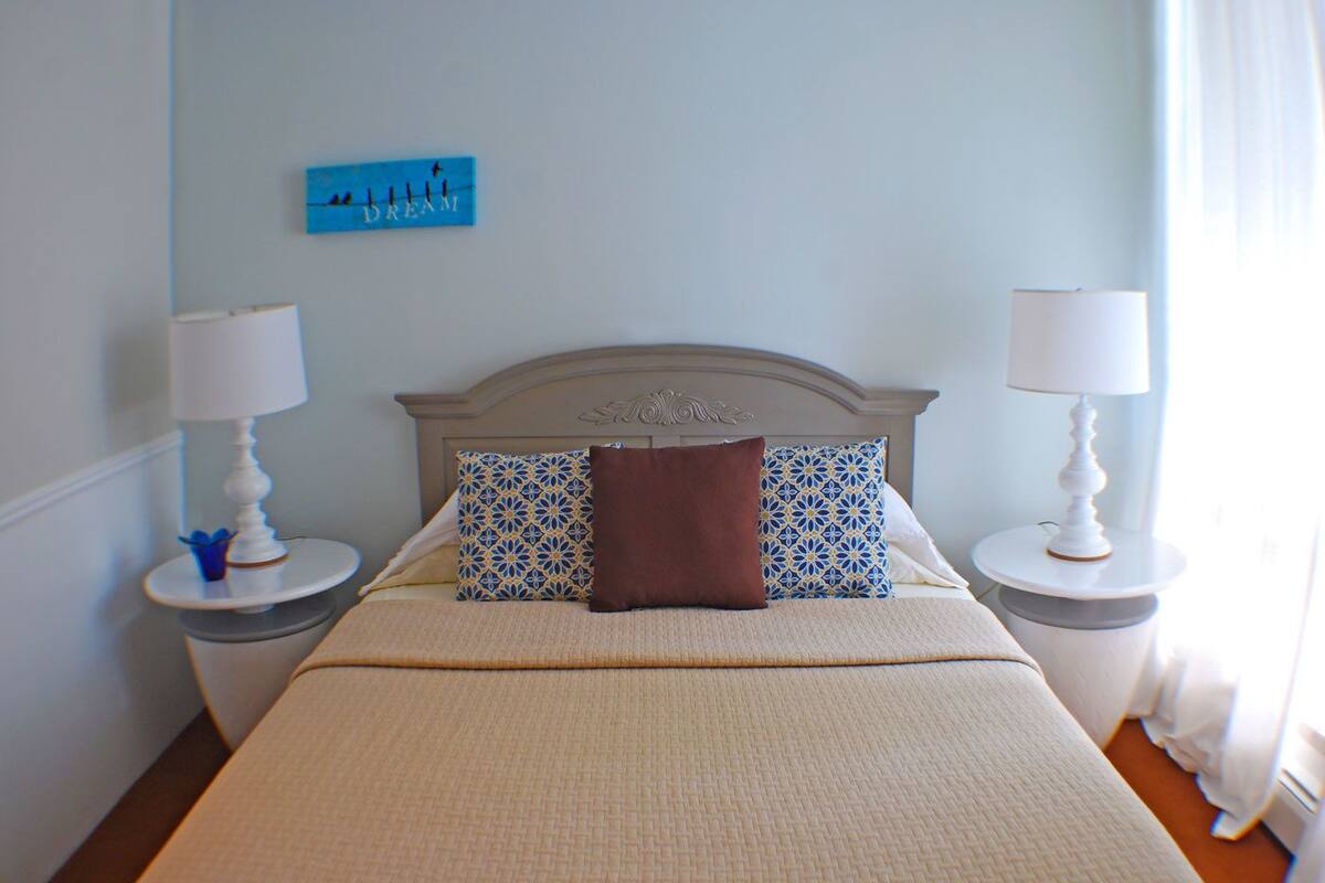 A neatly made bed with a patterned pillow, two bedside tables with lamps, and a blue artwork on the wall.