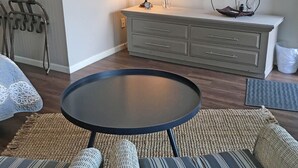 Una sala de estar con una mesa redonda, sofá de mimbre, un televisor montado en la pared y un mueble con cajones.