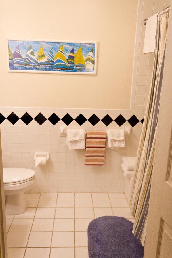 A bathroom with a toilet, a shower, a towel rack with towels, and a painting of sailboats on the wall.