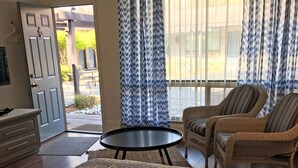 Una habitación con una cama, dos sillones de mimbre, una mesita redonda, un aparador y una puerta que da al exterior.
