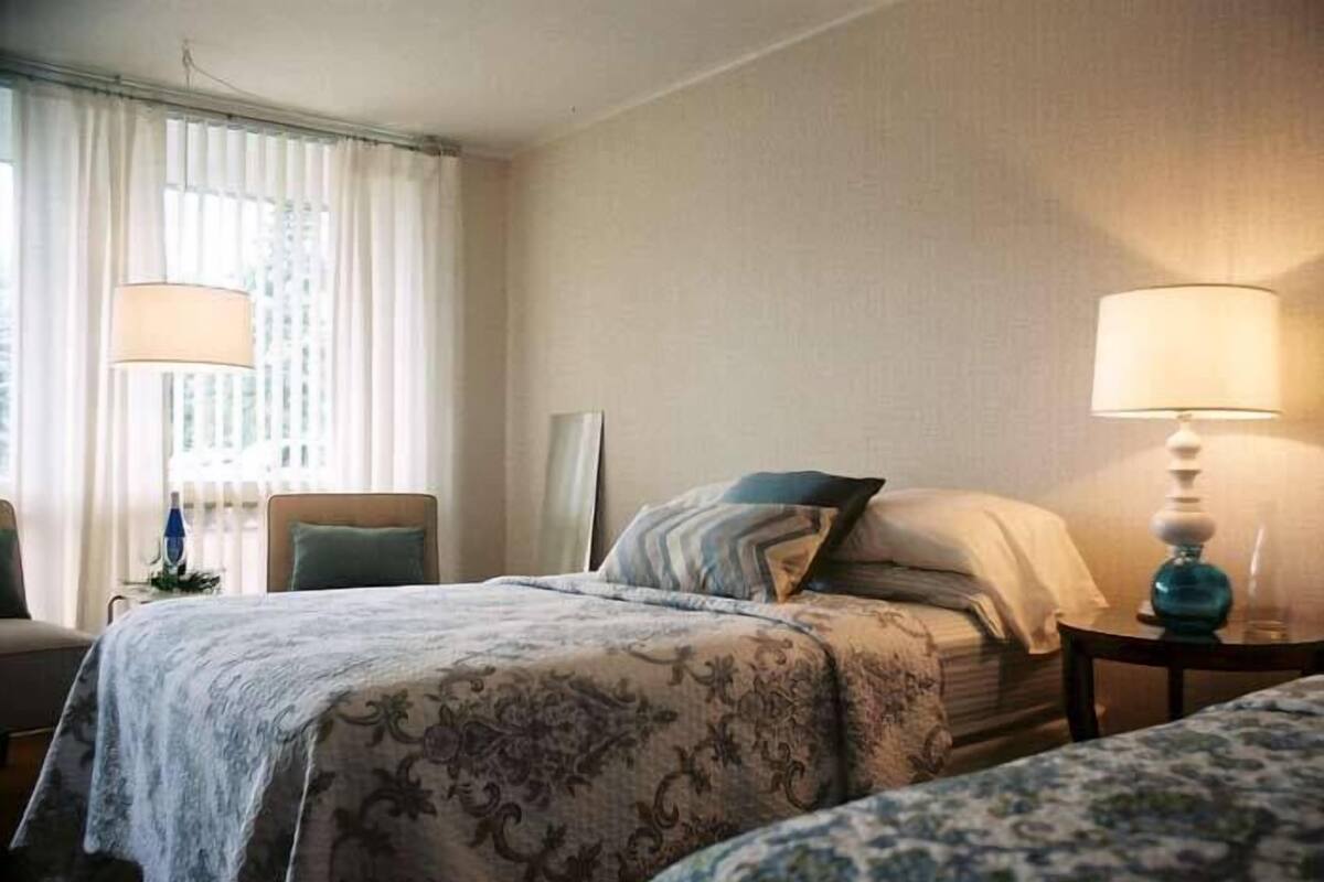 A hotel room with a bed, two bedside lamps, a chair, and a window with curtains.