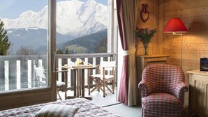 Una habitación con una cama, un sillón escocés, una mesita con sillas, una lámpara roja y vistas a montañas nevadas.