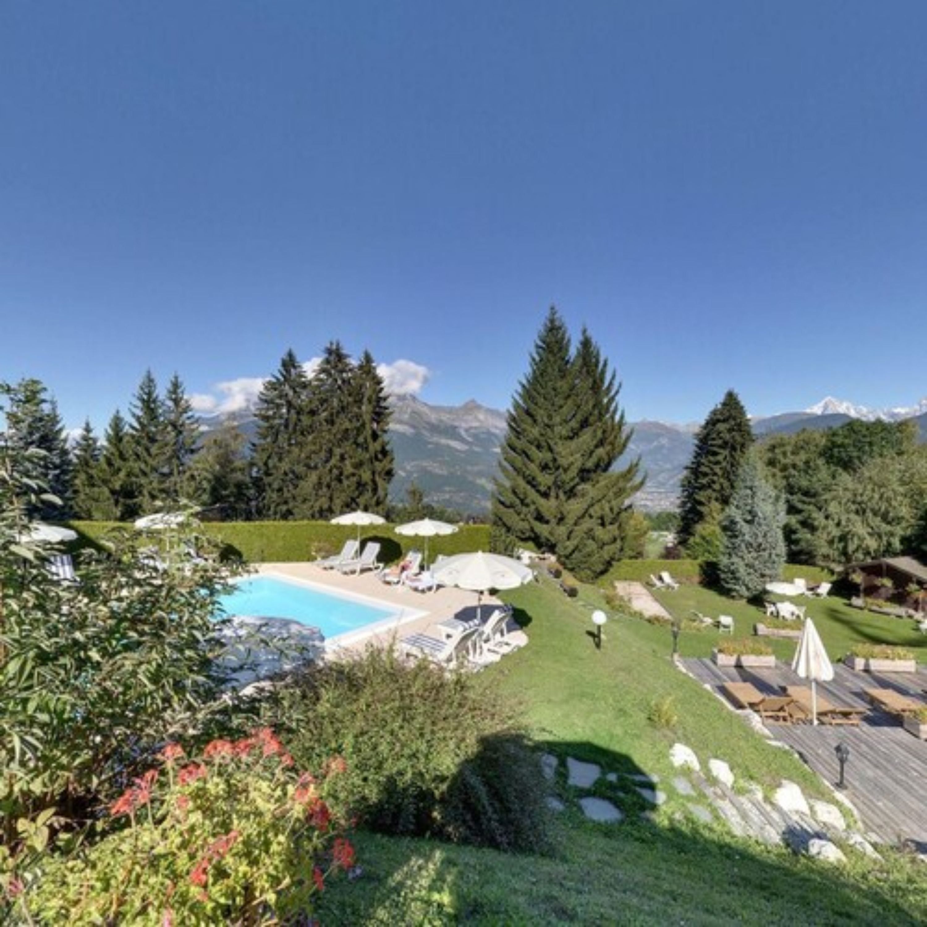 A pool area with lounge chairs and umbrellas, surrounded by greenery and mountains in the background.