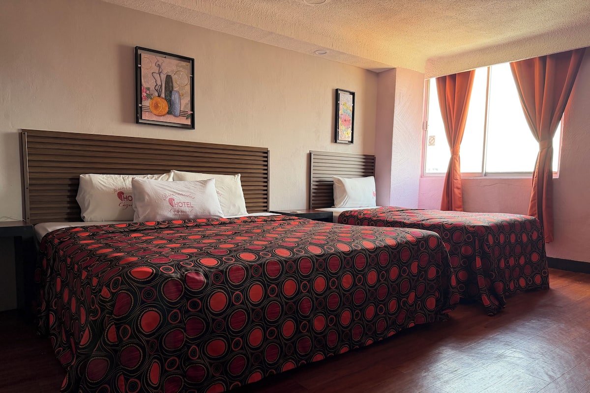 Een hotelkamer met twee bedden, houten hoofdeinden en een groot raam met gordijnen.