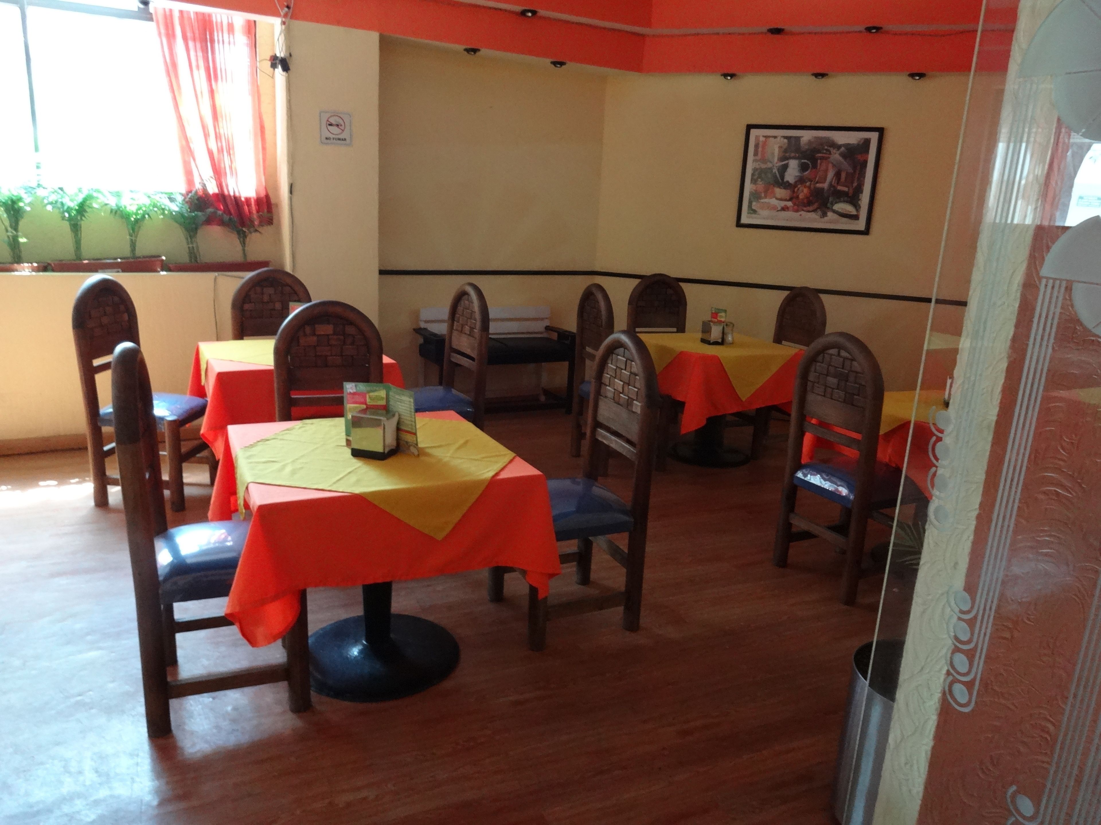Een restaurant met houten stoelen en tafels, sommige met gele en rode tafelkleden. Er staan planten op de vensterbank en een lijst aan de muur.