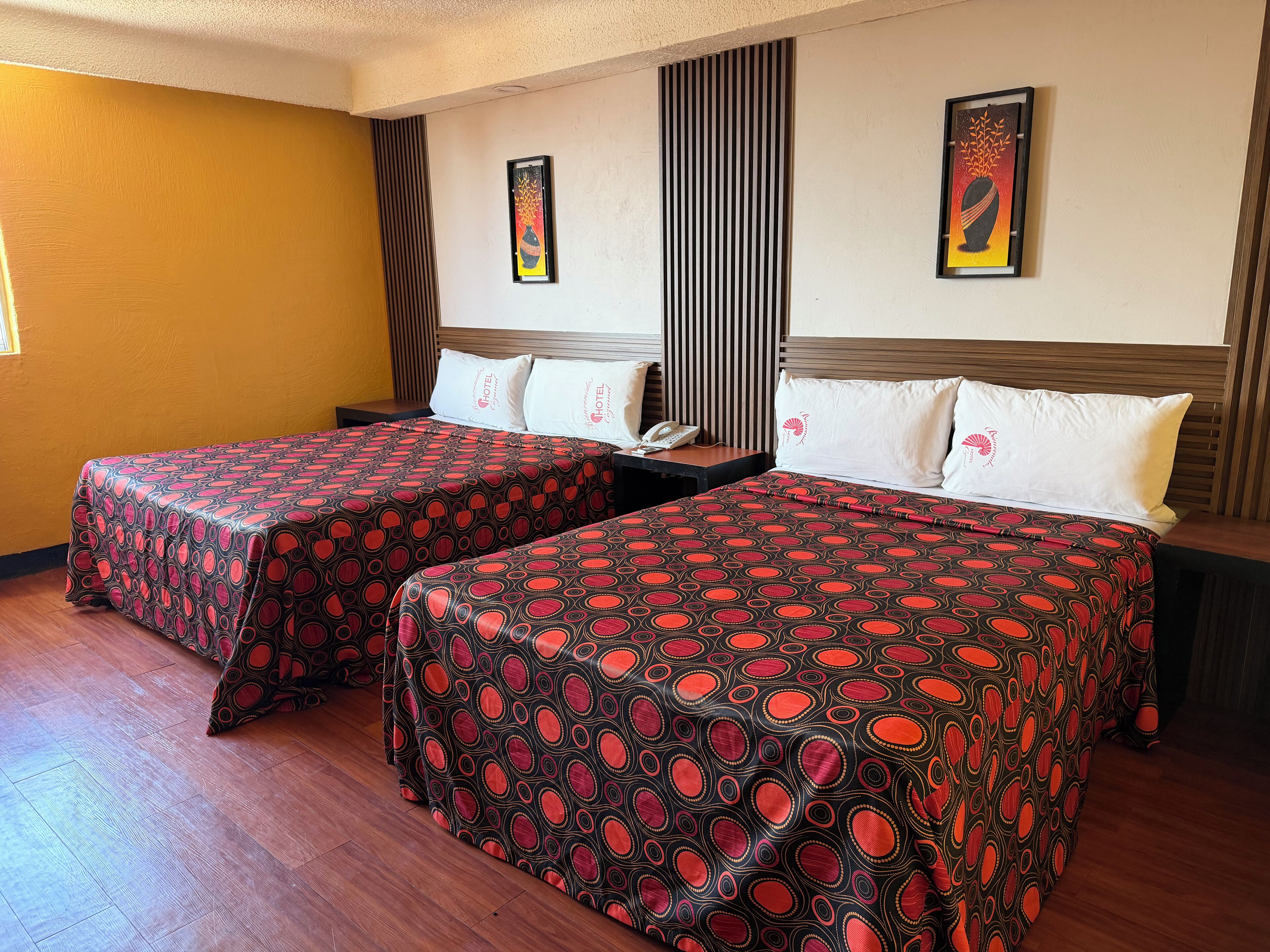 A hotel room with two beds, a wooden floor, a window with blinds, and artwork on the walls.