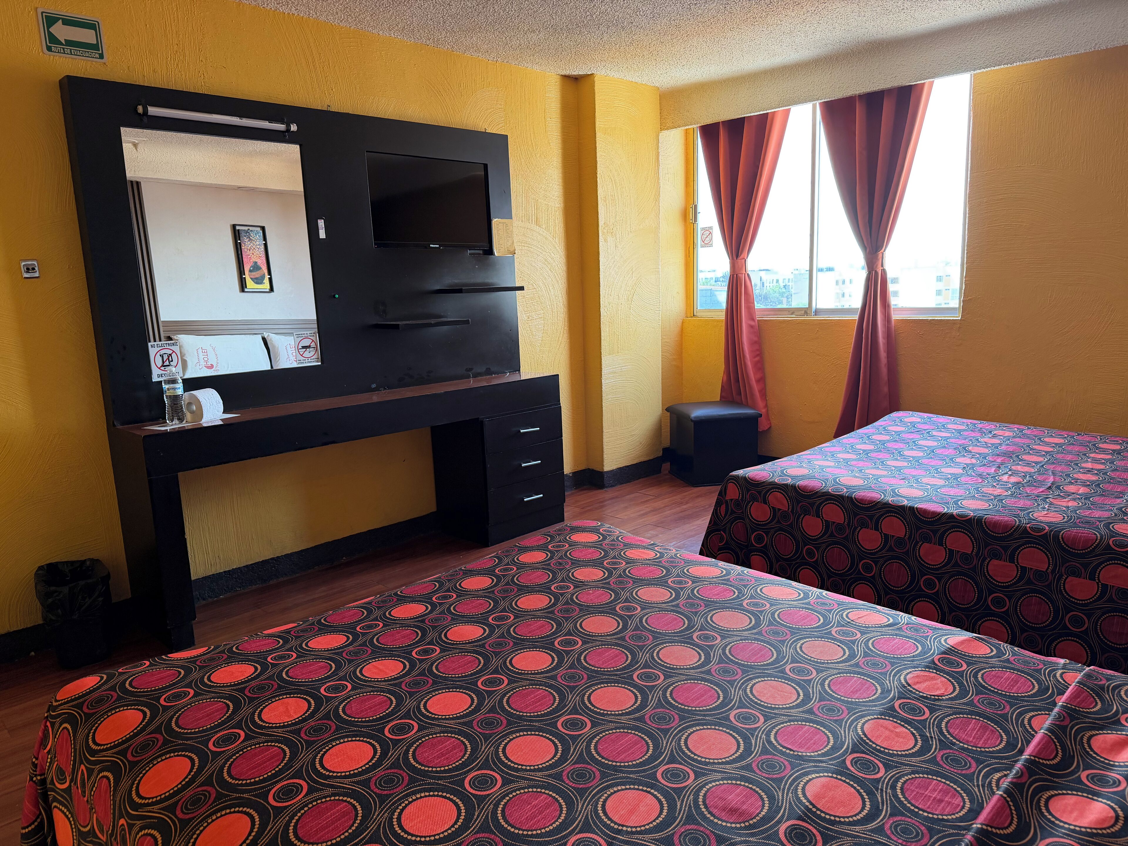 A hotel room with a desk, a television, two beds with patterned bedspreads, a window with curtains, and a view of the city.