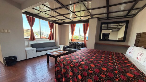 Habitación de hotel con una cama grande, un sofá, una mesita y vistas a la ciudad a través de amplios ventanales.