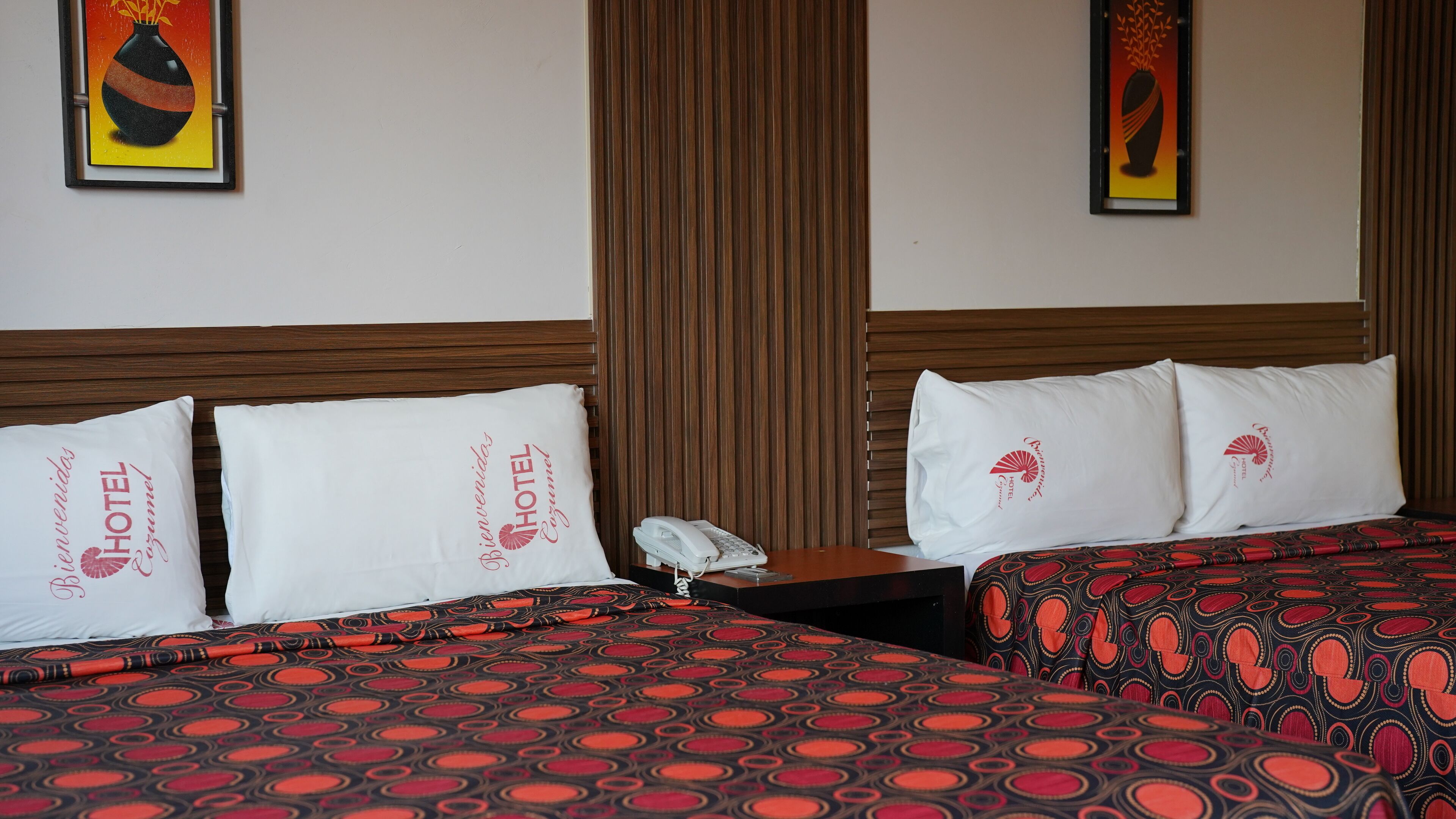 A hotel room with two beds, each with a patterned bedspread, white pillows with the hotel logo, a bedside table with a phone, and a wooden headboard.