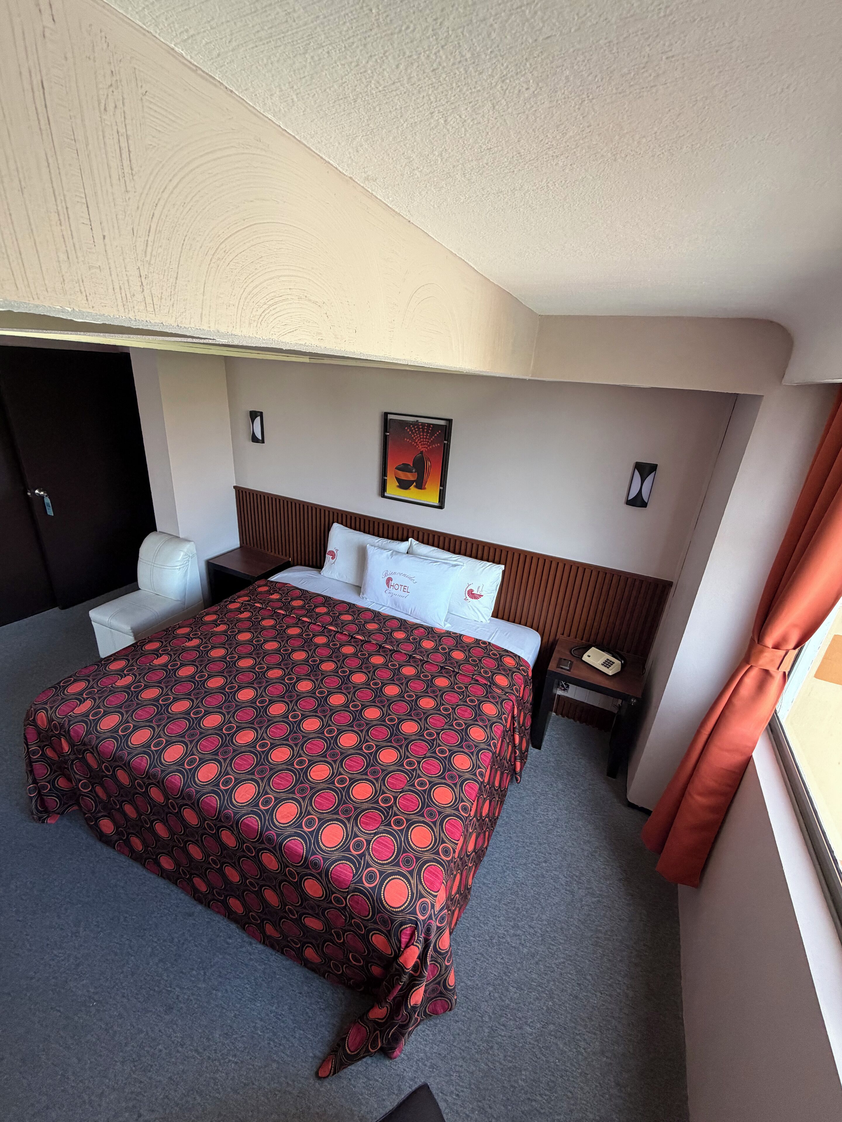 A hotel room with a bed, a desk, a chair, a nightstand, a telephone, and a window with curtains.