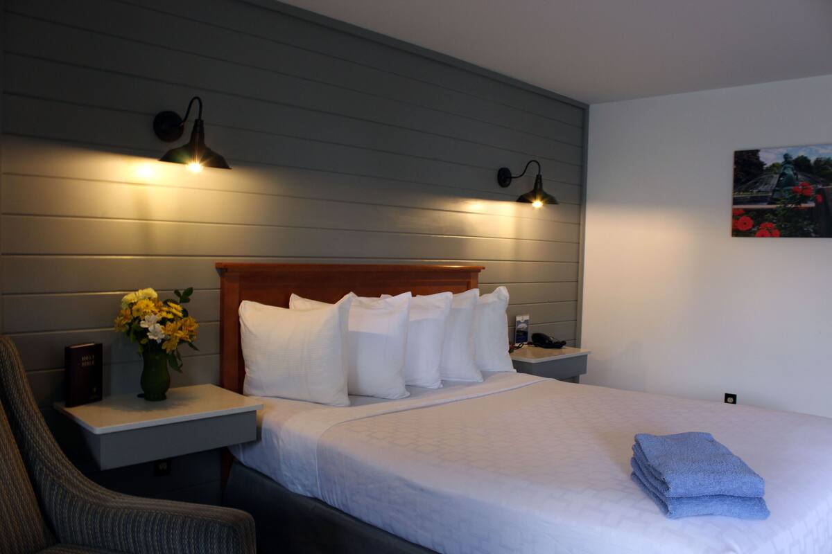 A hotel room with a neatly made bed, a chair, a nightstand with a lamp, and a painting on the wall.