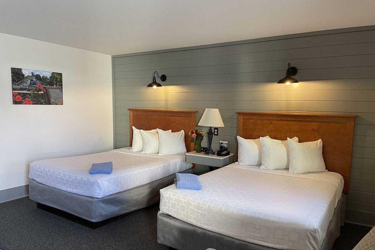 A hotel room with two single beds, a nightstand with a lamp, a small table with a vase of flowers, and a painting on the wall.