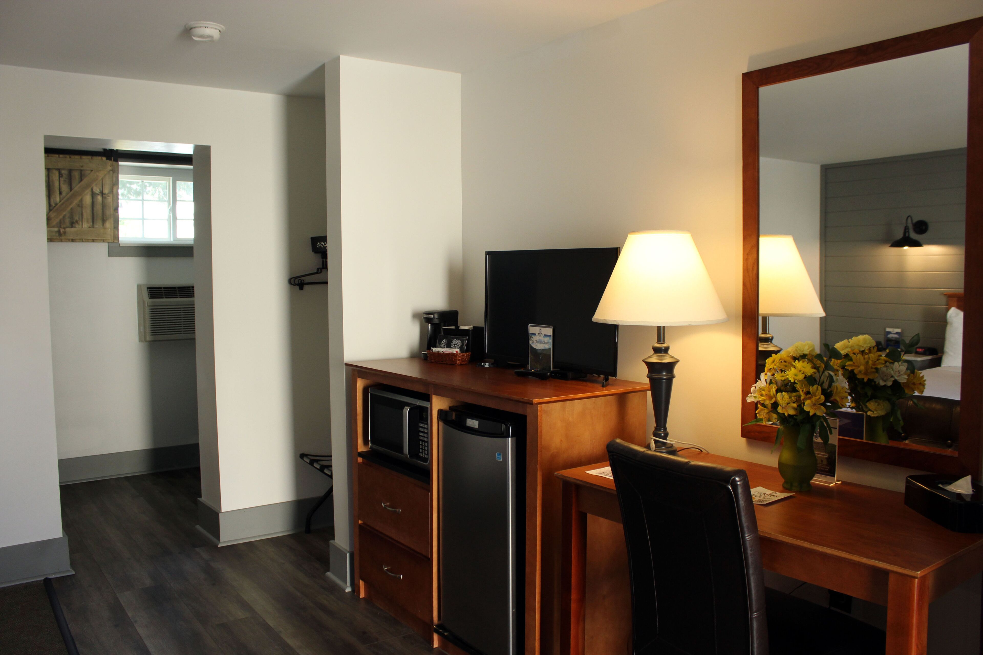 A compact hotel room with a kitchenette, a flat-screen TV, a desk with a lamp, and a mirror.
