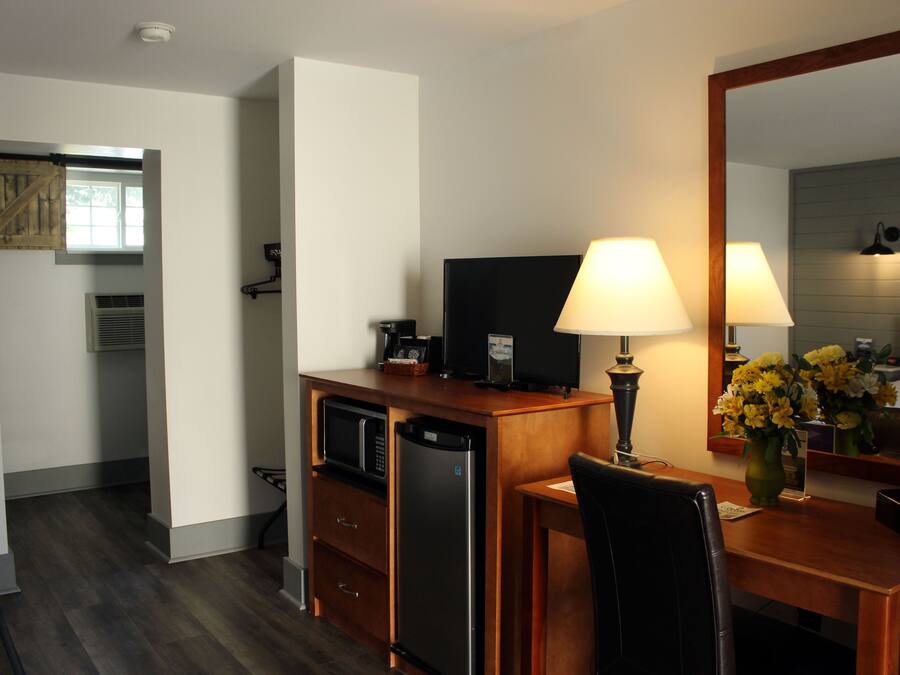 A compact hotel room with a kitchenette, a flat-screen TV, a desk with a lamp, and a mirror.