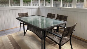 A covered patio with a glass table and wicker chairs, surrounded by white wooden walls and a view of trees. - Parkside Cottages and Suites (Stanhope)