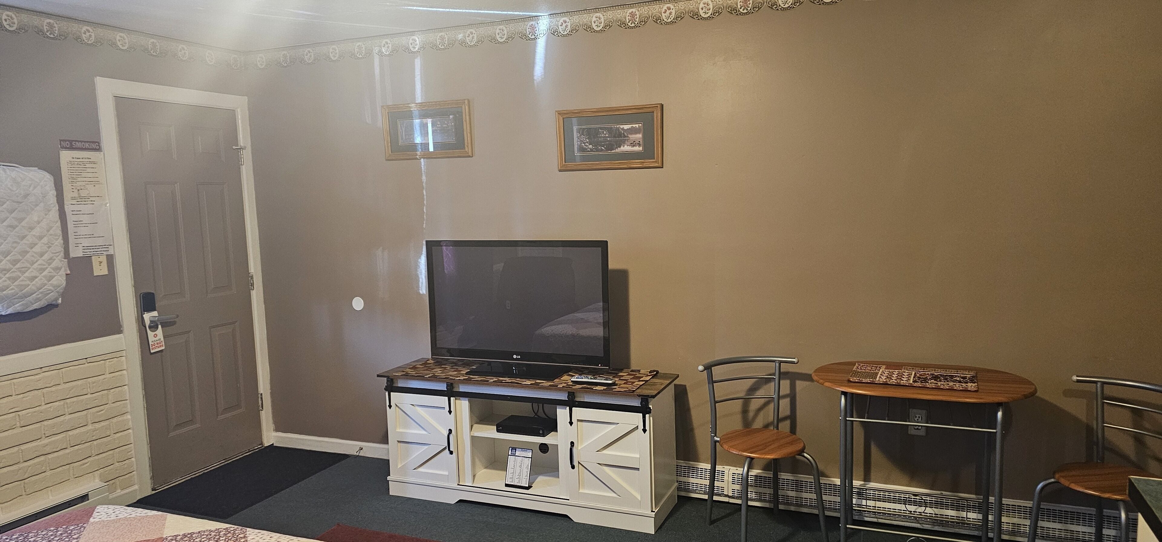 A room with a television on a wooden cabinet, two chairs, a small table, a door with a 'No Smoking' sign, and two framed pictures on the wall.