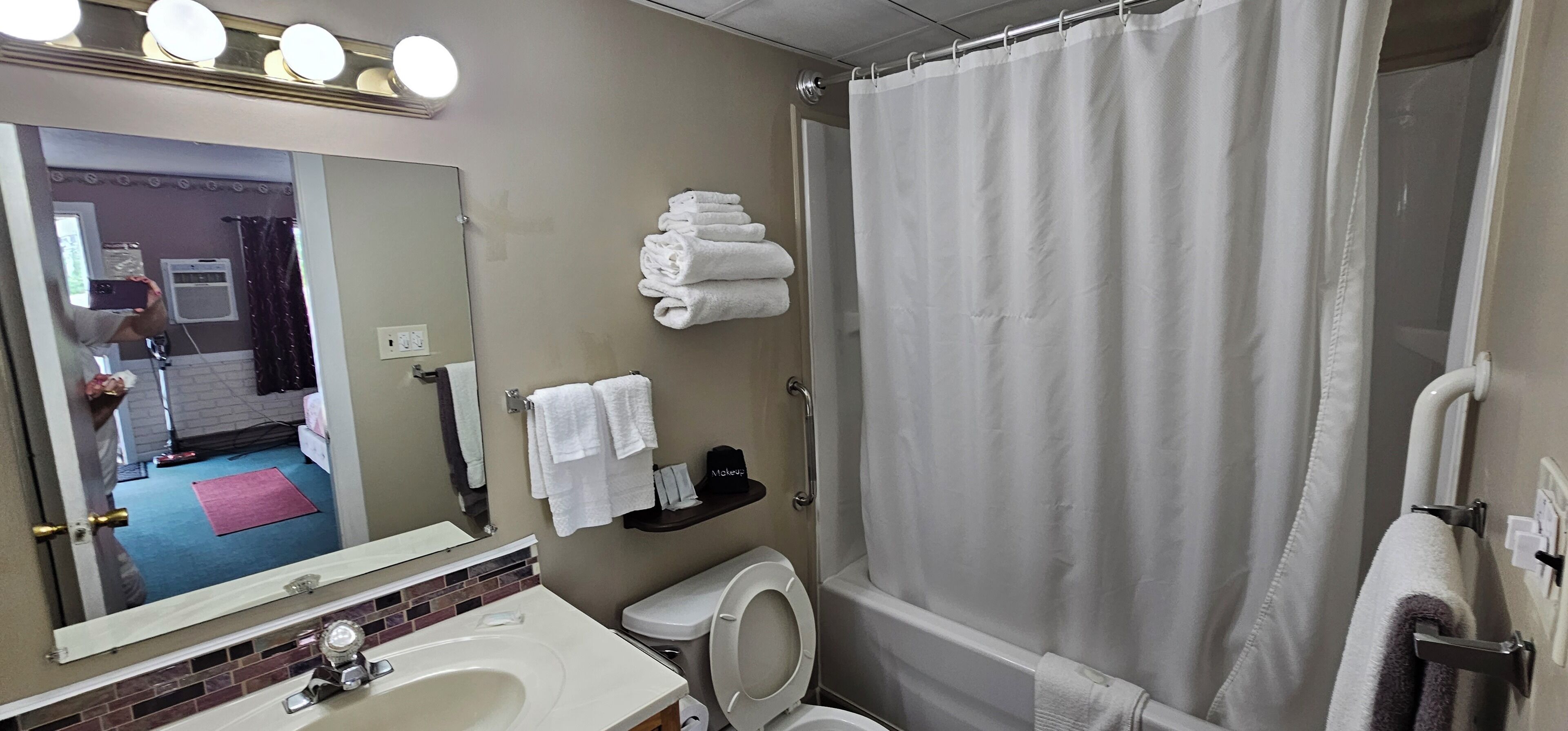Une salle de bain avec un lavabo, des toilettes et un rideau de douche. Des serviettes sont pliées sur une étagère et un miroir est accroché au-dessus du lavabo.