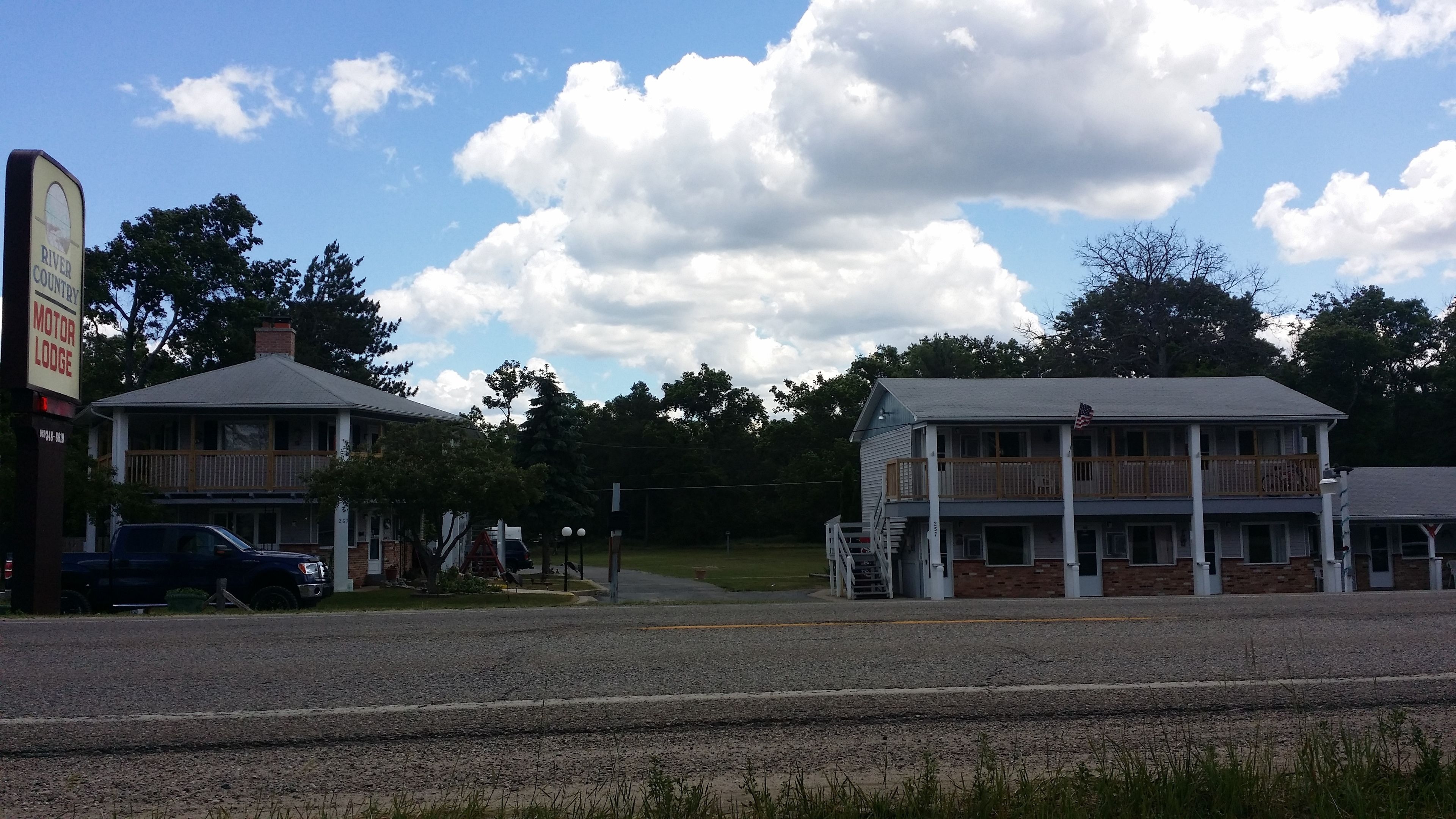 A two-story building with a porch and a flag, likely a lodging establishment, with a road in the foreground and trees in the background.
