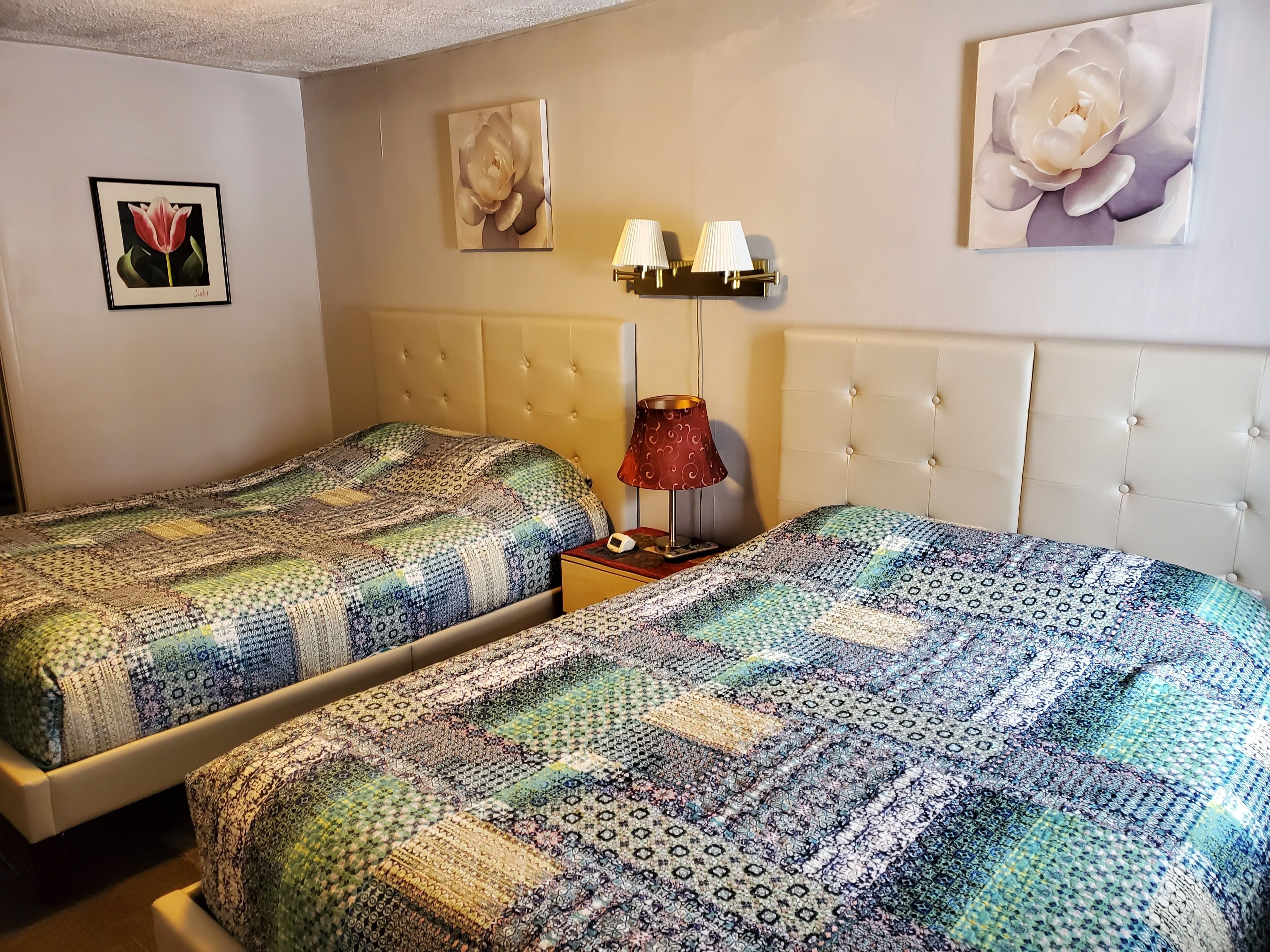 A hotel room with two beds, a tufted headboard, wall-mounted lamps, and framed floral artwork.