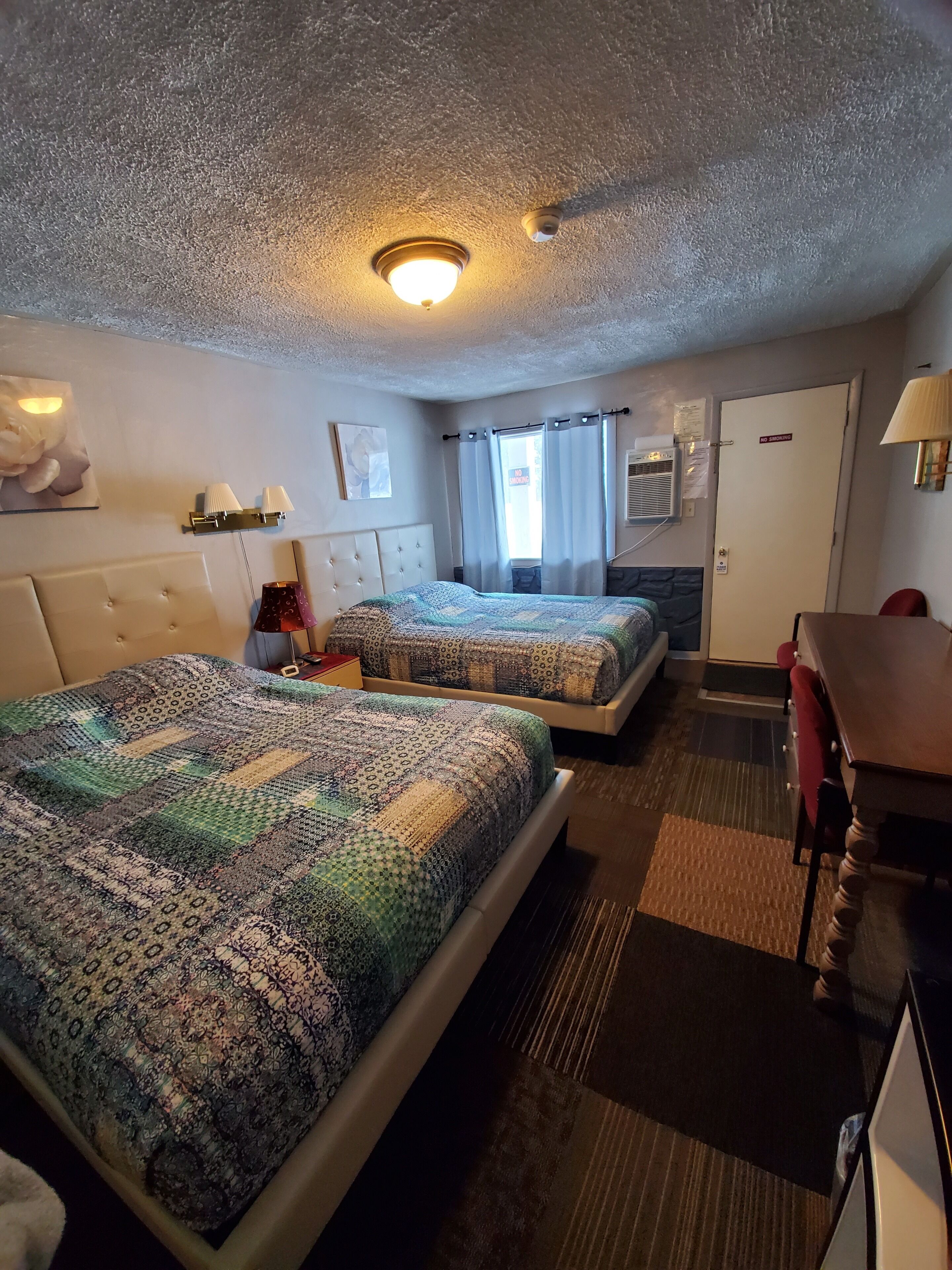 A hotel room with two beds, a desk, a chair, a lamp, a ceiling light, and a door with a 'No Smoking' sign.