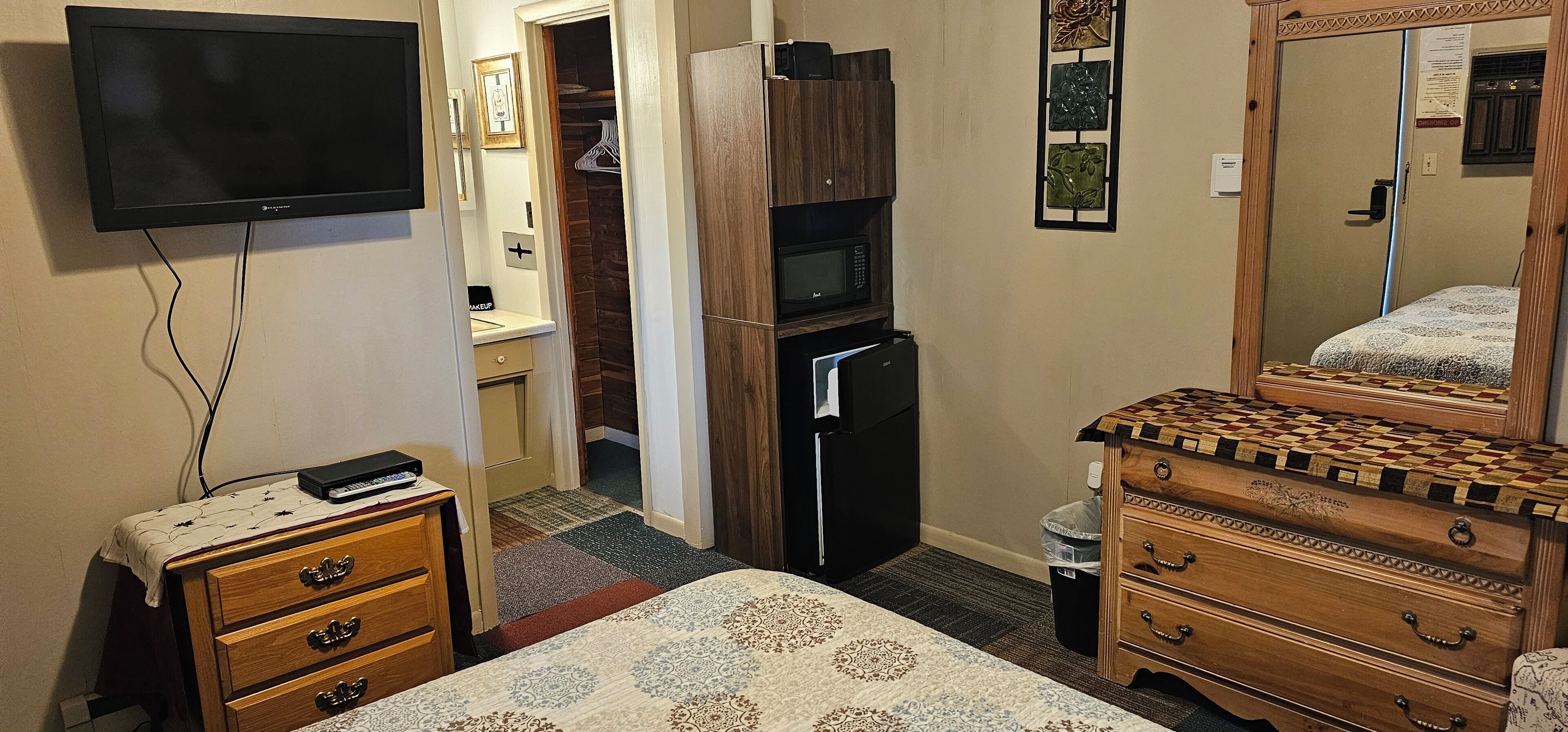 Ein kompaktes Hotelzimmer mit einem an der Wand montierten Flachbildfernseher, einer Holzkommode mit kariertem Deckel, einem Bett mit gemustertem Bettwäsche und einem Kühlschrank.