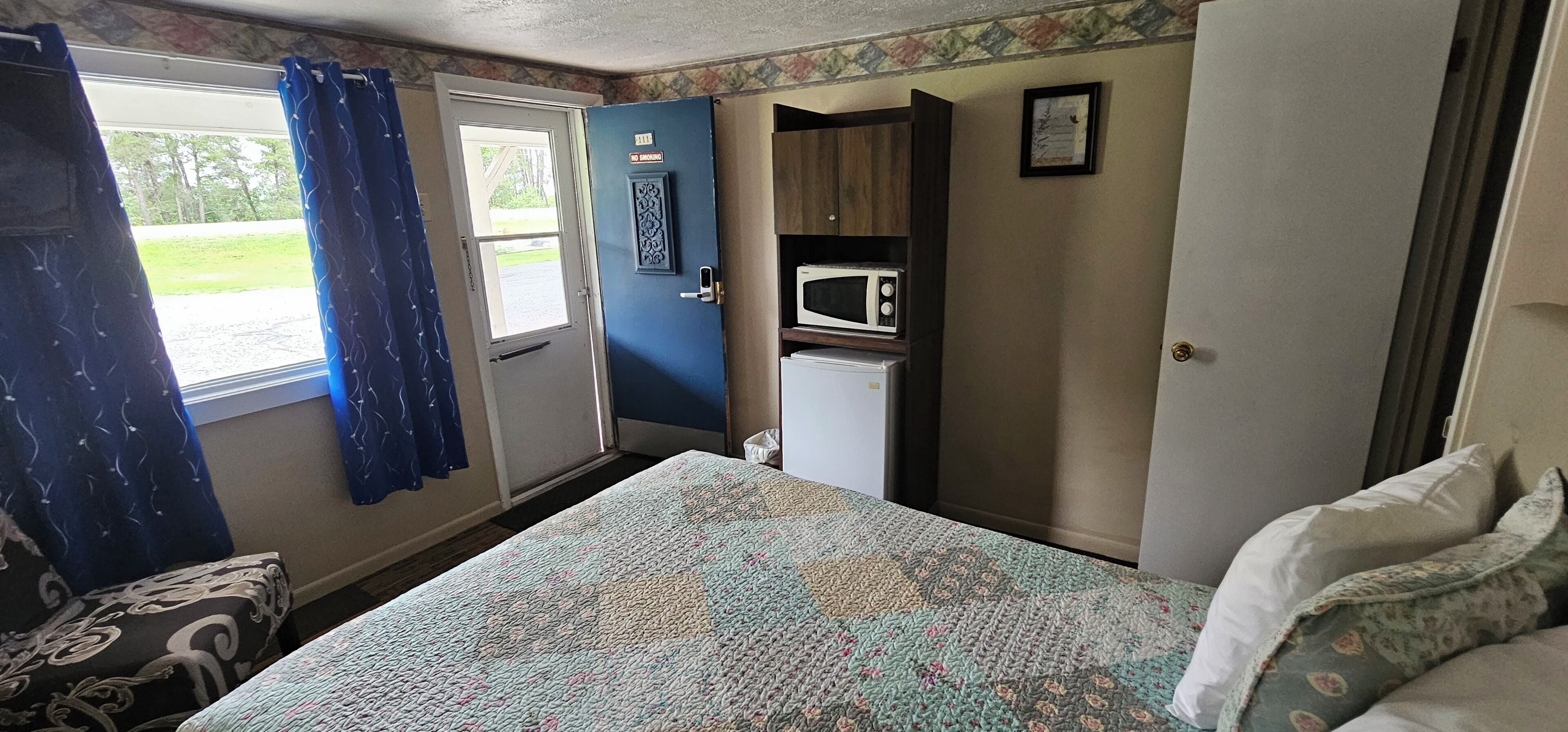 Une chambre de taille modeste comprenant un lit, une petite kitchenette équipée d’un four à micro-ondes, une porte sur laquelle est apposé un panneau « Interdiction de fumer » et une fenêtre avec des rideaux.