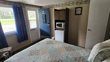 Une chambre de taille modeste comprenant un lit, une petite kitchenette équipée d’un four à micro-ondes, une porte sur laquelle est apposé un panneau « Interdiction de fumer » et une fenêtre avec des rideaux.