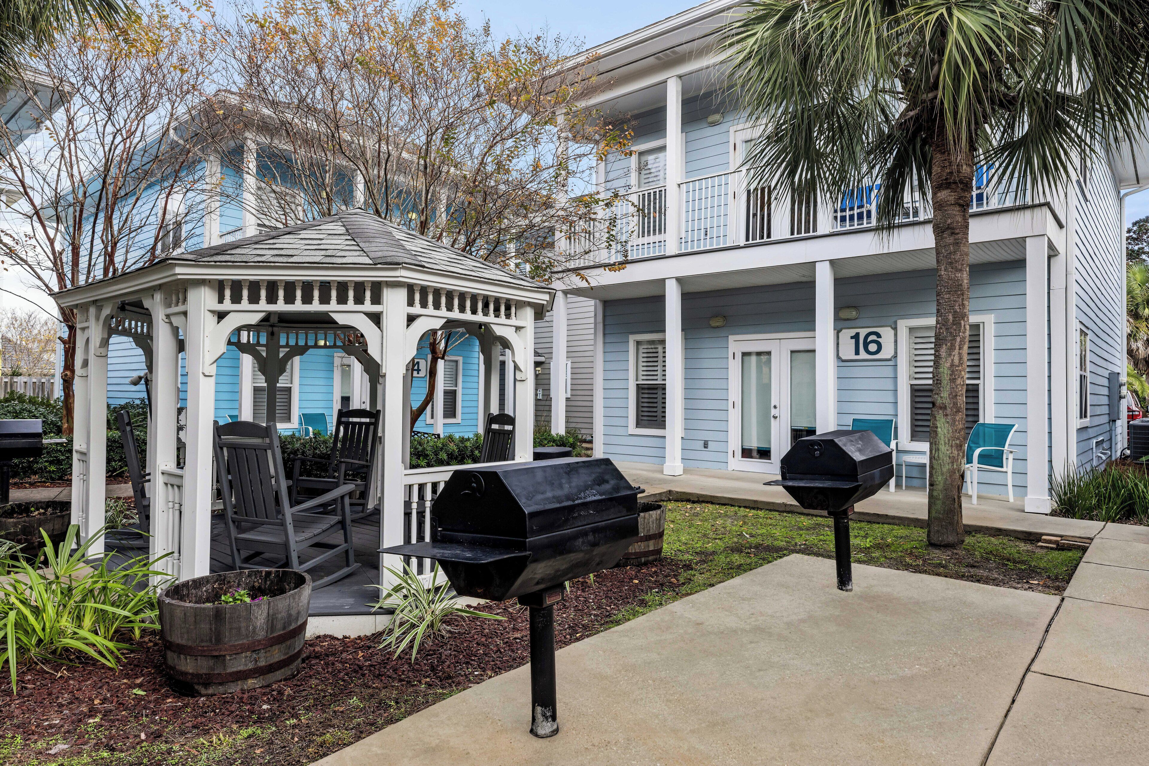 A gazebo with chairs, a barbecue grill, and a mailbox in a landscaped area with a building in the background.