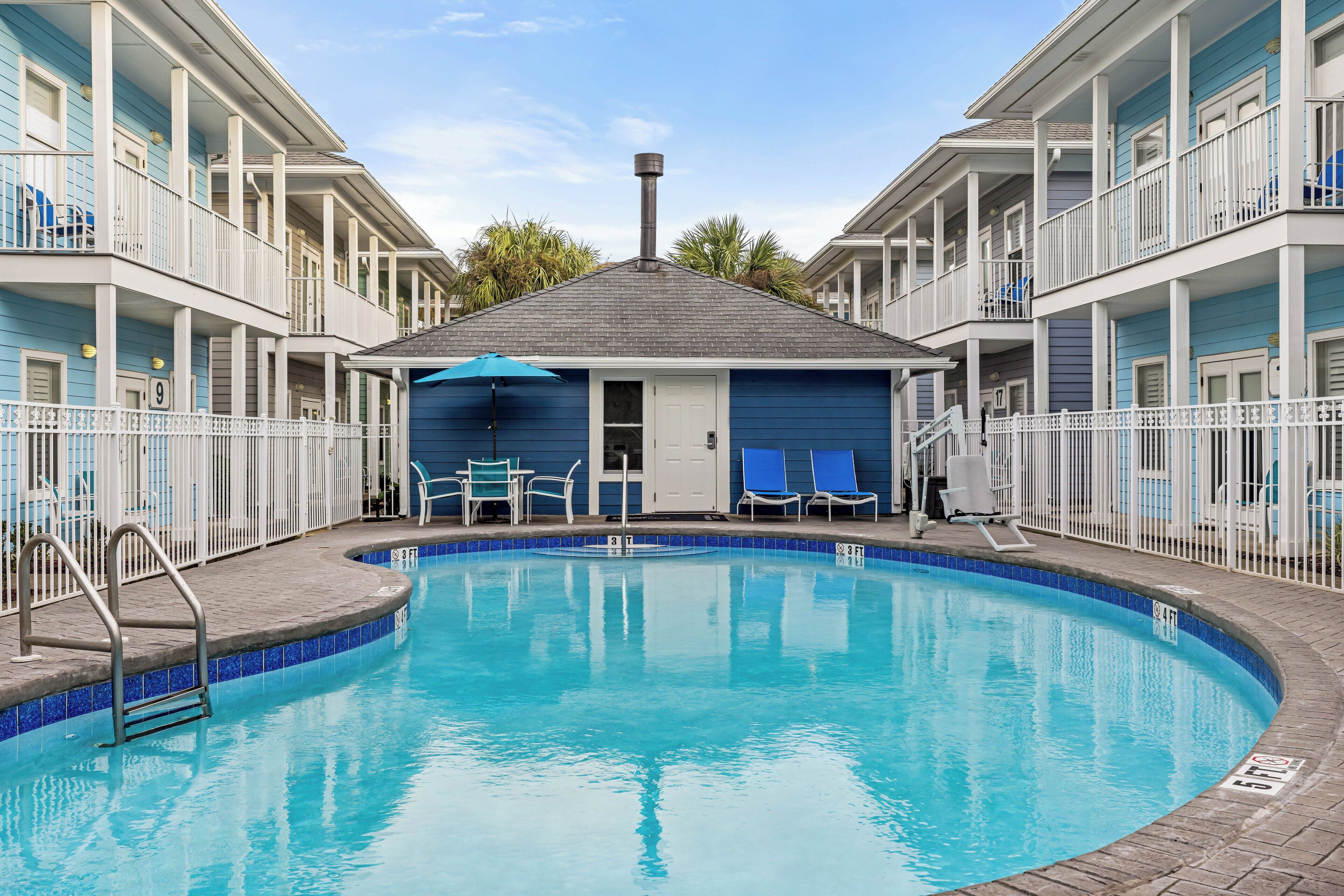 A swimming pool surrounded by a white fence, with a building featuring balconies and a door in the center.