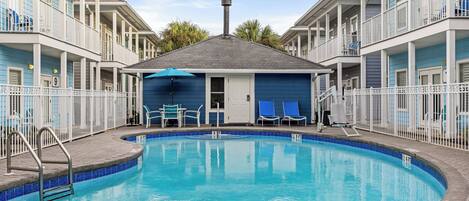 A swimming pool surrounded by a white fence, with a building featuring balconies and a door in the center.