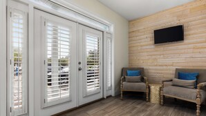 Quarto com porta de vidro deslizante, televisão de tela plana fixada em parede de madeira, duas poltronas com almofadas azuis e uma pequena mesa redonda.