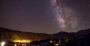 A night sky with stars and the Milky Way over a forested landscape with a lake and illuminated buildings. - June Lake Motel (June Lake)