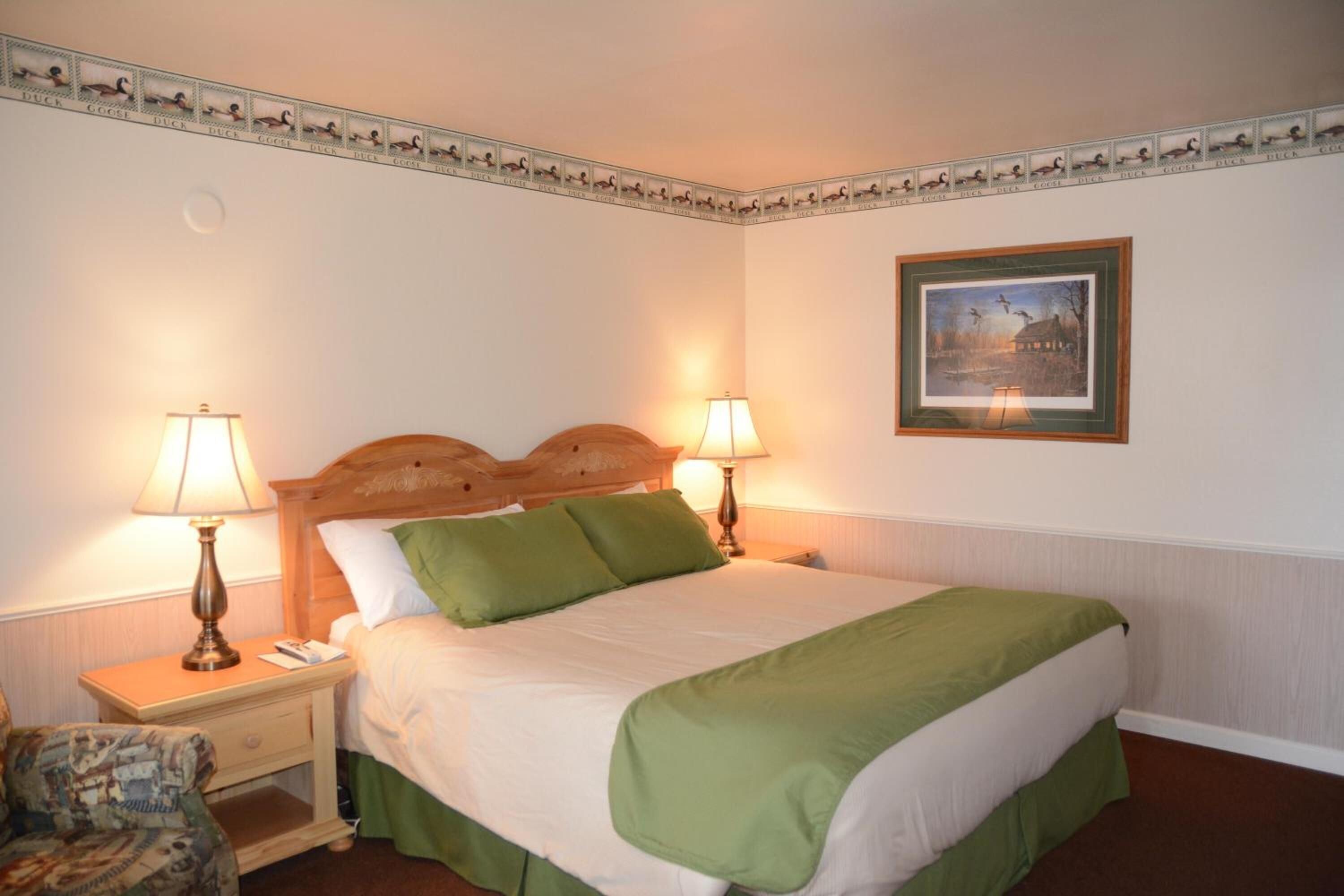 Habitación de hotel con una cama grande, dos lámparas de noche, un cuadro enmarcado en la pared y un borde decorativo sobre la cama.
