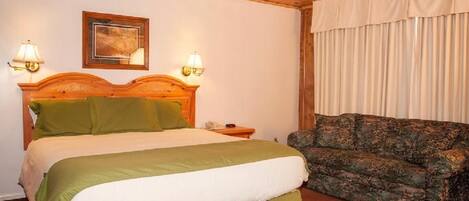 A room with a bed, two bedside lamps, a wooden headboard, a framed picture on the wall, and a patterned sofa.