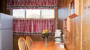 A wooden cabin interior with a dining table, chairs, a refrigerator, and a window with checkered curtains. - June Lake Motel (June Lake)