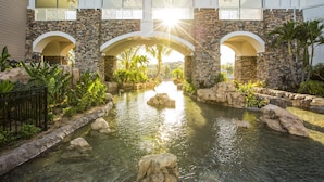 Un pont en pierre, doté d’arches enjambant un bassin d’eau, entouré de plantes tropicales et de palmiers.