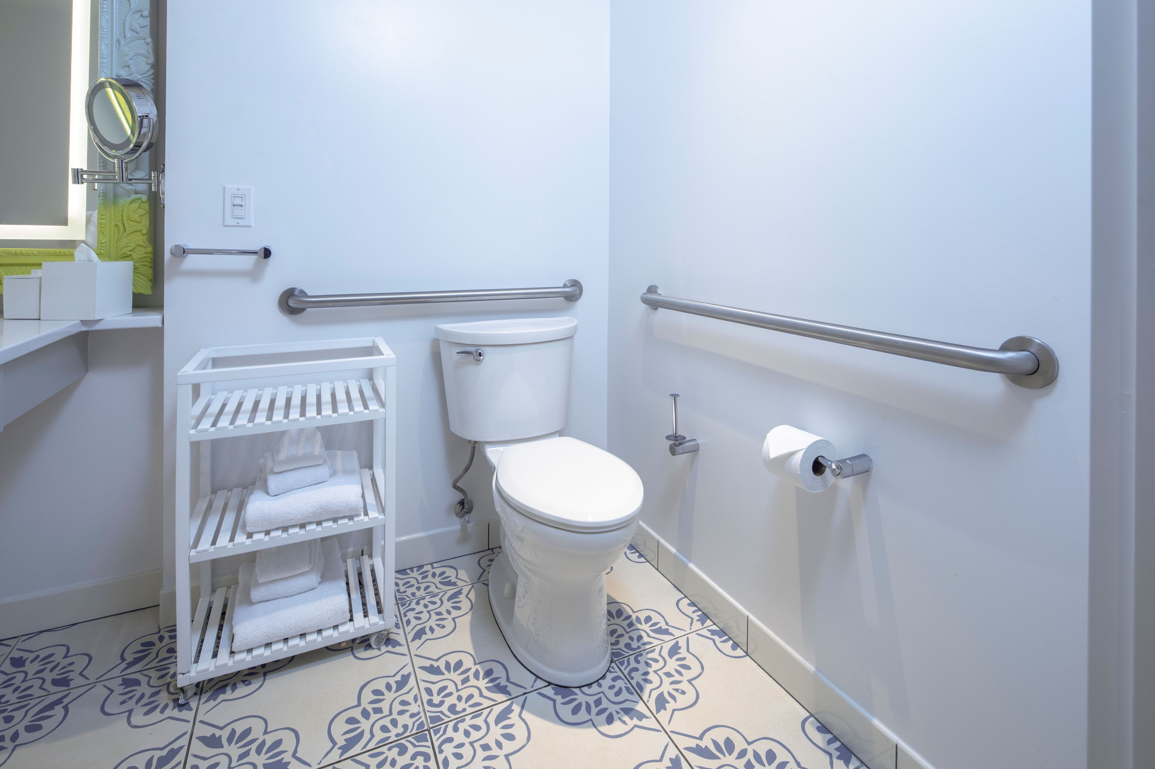 A bathroom with a toilet, handrails, a shelf with towels, and patterned floor tiles.
