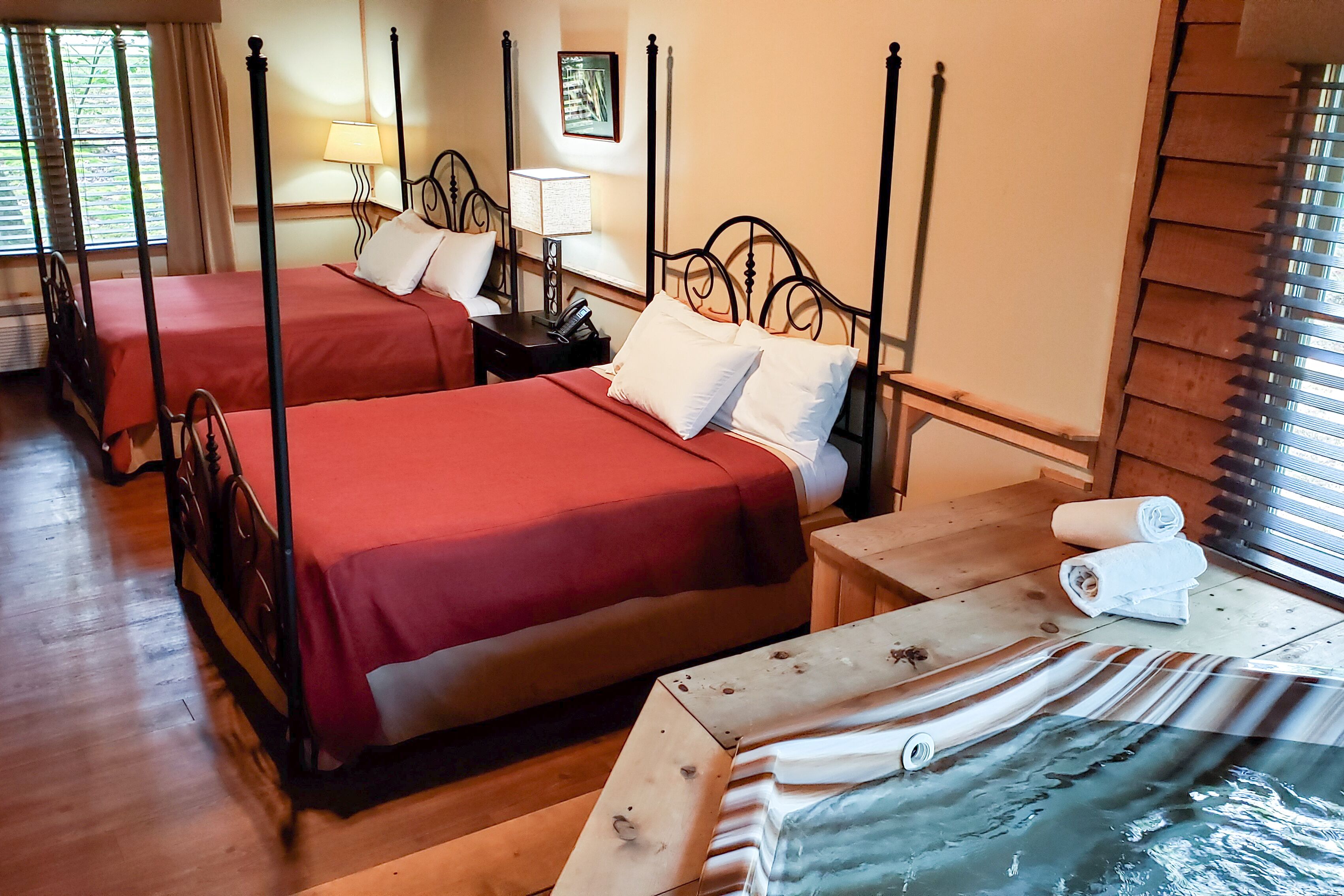 A hotel room with two four-poster beds, wooden flooring, a wooden nightstand with a lamp, and a window with blinds.