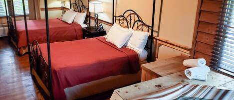 A hotel room with two four-poster beds, wooden flooring, a wooden nightstand with a lamp, and a window with blinds.