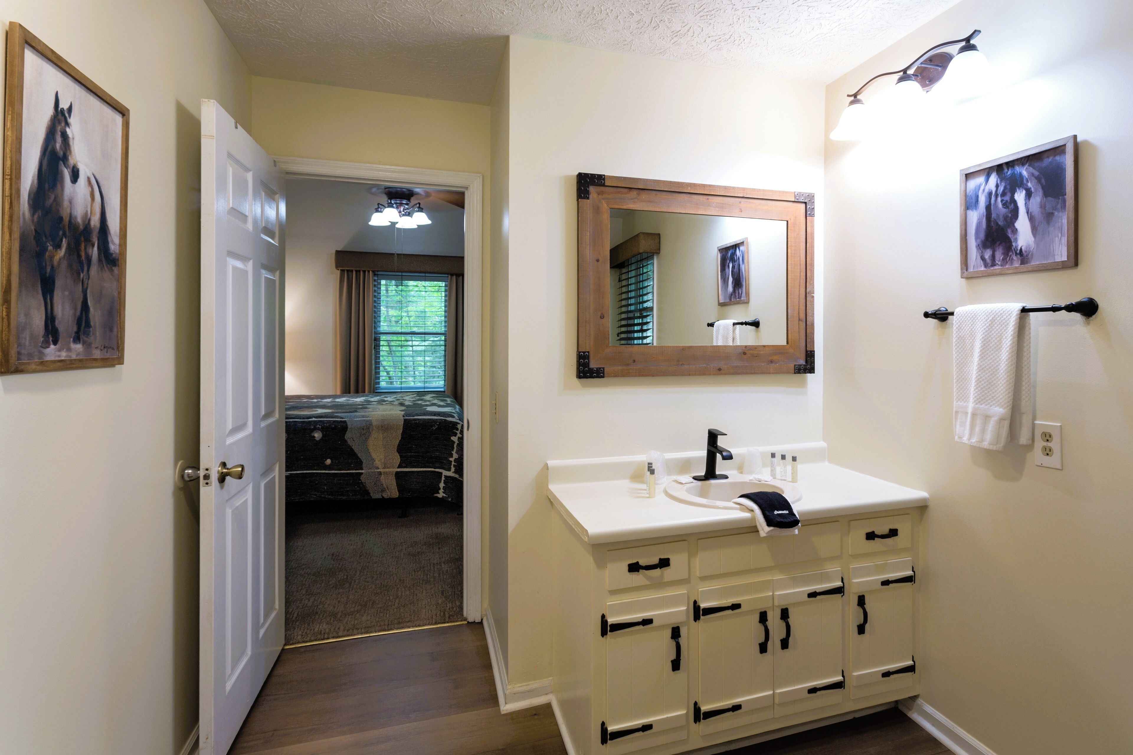 A bathroom with a white sink, mirror, and cabinets. There is a door leading to a bedroom with a bed and a window.