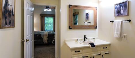 A bathroom with a white sink, mirror, and cabinets. There is a door leading to a bedroom with a bed and a window.