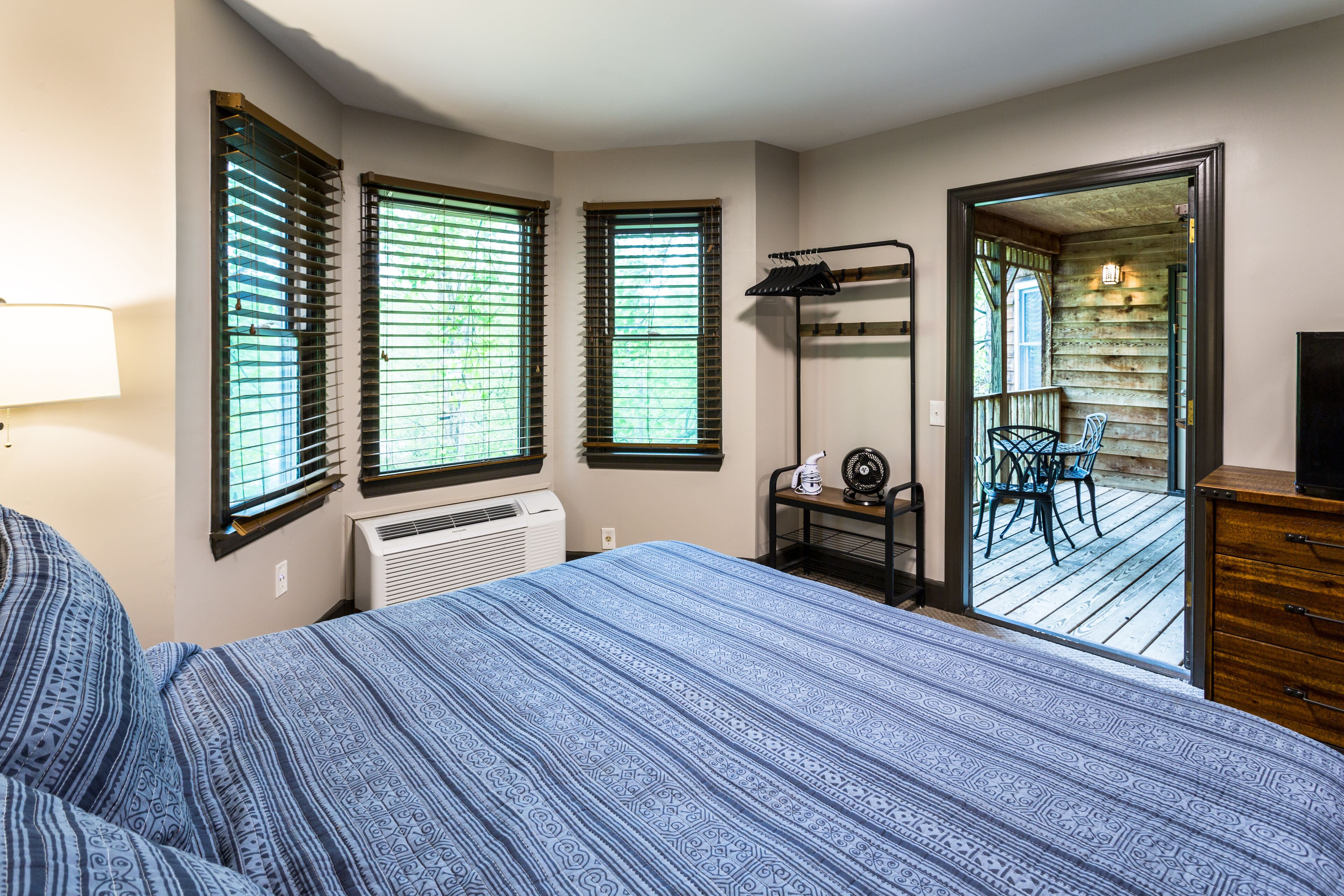 A bedroom with a bed, two windows with blinds, a heater, a TV, and a view of an outdoor deck.