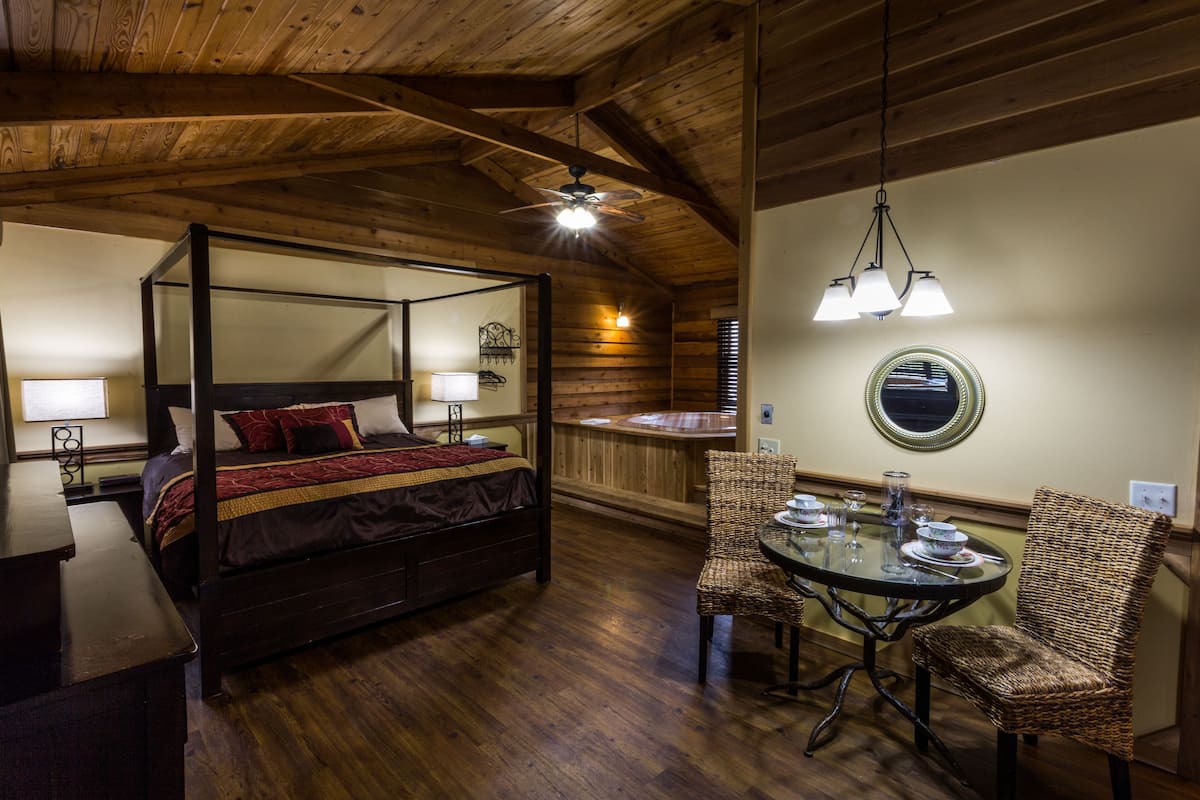 A four-poster bed with a red and gold bedspread, a dining table with chairs, and a ceiling fan.